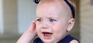 A baby girl wearing a navy dress and headband cries while tugging at her ear because of an incoming tooth